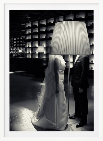 A surreal black and white photograph of a bride and groom whose heads are comically obscured by a large, pleated lampshade from a floor lamp standing between them. They are in a modern, dimly lit room with a wall of illuminated shelves. Poster
