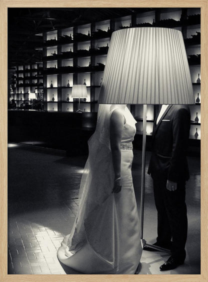 A surreal black and white photograph of a bride and groom whose heads are comically obscured by a large, pleated lampshade from a floor lamp standing between them. They are in a modern, dimly lit room with a wall of illuminated shelves. Poster