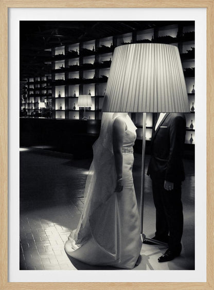 A surreal black and white photograph of a bride and groom whose heads are comically obscured by a large, pleated lampshade from a floor lamp standing between them. They are in a modern, dimly lit room with a wall of illuminated shelves. Poster