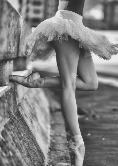 A black and white photograph focusing on the lower half of a ballerina in a tutu and pointe shoes. She stands en pointe with one foot on the ground, while the other foot rests against a rough, concrete curb, highlighting the contrast between delicate dance and an urban environment. Decor