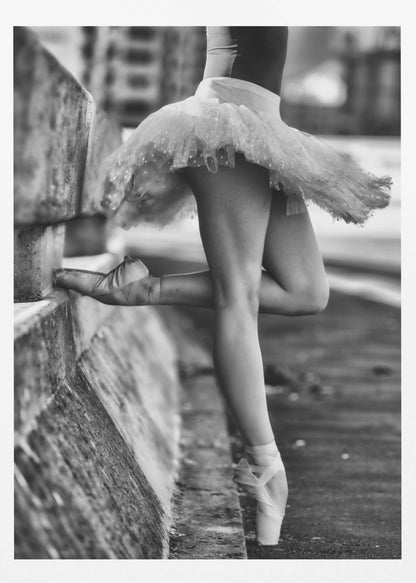 A black and white photograph focusing on the lower half of a ballerina in a tutu and pointe shoes. She stands en pointe with one foot on the ground, while the other foot rests against a rough, concrete curb, highlighting the contrast between delicate dance and an urban environment. Decor