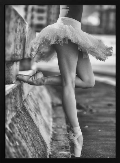 A black and white photograph focusing on the lower half of a ballerina in a tutu and pointe shoes. She stands en pointe with one foot on the ground, while the other foot rests against a rough, concrete curb, highlighting the contrast between delicate dance and an urban environment. Decor