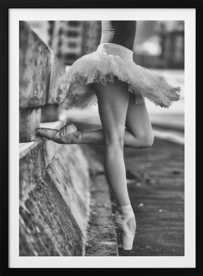 A black and white photograph focusing on the lower half of a ballerina in a tutu and pointe shoes. She stands en pointe with one foot on the ground, while the other foot rests against a rough, concrete curb, highlighting the contrast between delicate dance and an urban environment. Decor