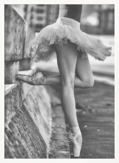 A black and white photograph focusing on the lower half of a ballerina in a tutu and pointe shoes. She stands en pointe with one foot on the ground, while the other foot rests against a rough, concrete curb, highlighting the contrast between delicate dance and an urban environment. Decor