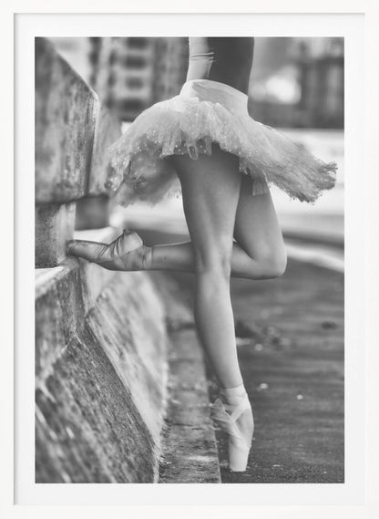 A black and white photograph focusing on the lower half of a ballerina in a tutu and pointe shoes. She stands en pointe with one foot on the ground, while the other foot rests against a rough, concrete curb, highlighting the contrast between delicate dance and an urban environment. Decor
