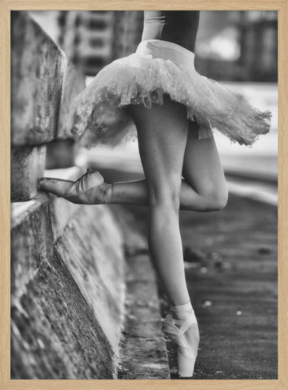 A black and white photograph focusing on the lower half of a ballerina in a tutu and pointe shoes. She stands en pointe with one foot on the ground, while the other foot rests against a rough, concrete curb, highlighting the contrast between delicate dance and an urban environment. Decor