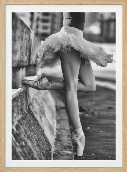 A black and white photograph focusing on the lower half of a ballerina in a tutu and pointe shoes. She stands en pointe with one foot on the ground, while the other foot rests against a rough, concrete curb, highlighting the contrast between delicate dance and an urban environment. Decor