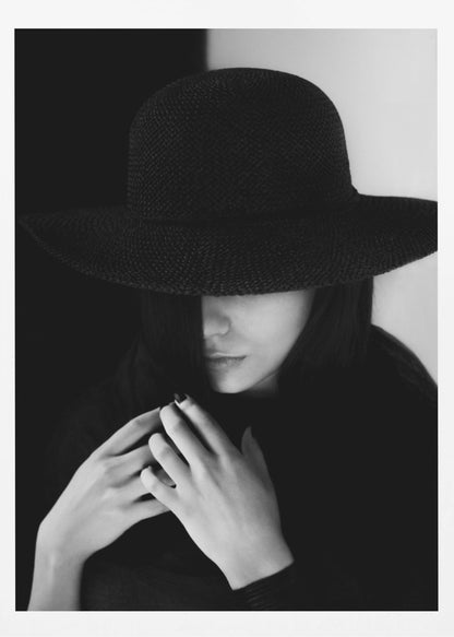 A mysterious black and white portrait of a woman wearing a large, black, wide-brimmed hat that casts a deep shadow over her eyes. Her hands are clasped at her chest, and the background is split vertically into black and white sections. Artwork
