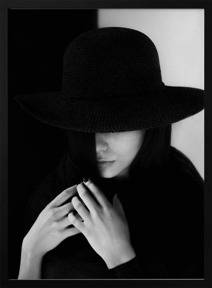 A mysterious black and white portrait of a woman wearing a large, black, wide-brimmed hat that casts a deep shadow over her eyes. Her hands are clasped at her chest, and the background is split vertically into black and white sections. Artwork