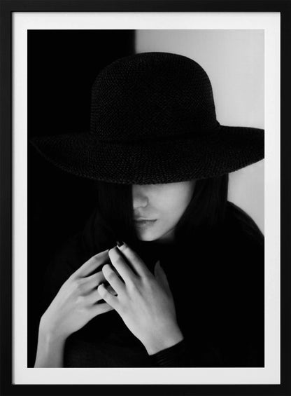 A mysterious black and white portrait of a woman wearing a large, black, wide-brimmed hat that casts a deep shadow over her eyes. Her hands are clasped at her chest, and the background is split vertically into black and white sections. Artwork