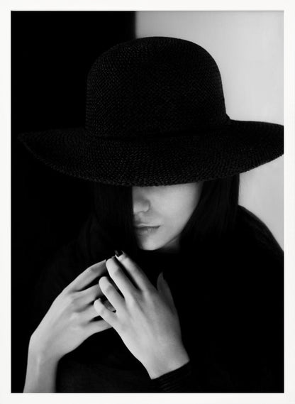A mysterious black and white portrait of a woman wearing a large, black, wide-brimmed hat that casts a deep shadow over her eyes. Her hands are clasped at her chest, and the background is split vertically into black and white sections. Artwork