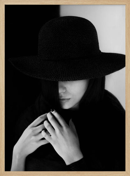A mysterious black and white portrait of a woman wearing a large, black, wide-brimmed hat that casts a deep shadow over her eyes. Her hands are clasped at her chest, and the background is split vertically into black and white sections. Artwork