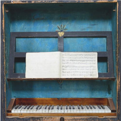 A vintage keyboard instrument is displayed inside a rustic wooden box with a distressed blue interior. An open book of sheet music rests on a black stand above the white and black keys. The entire piece is set within a simple, light-colored wood frame. Decor