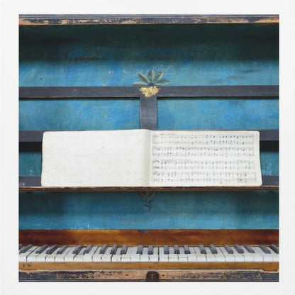 A vintage keyboard instrument is displayed inside a rustic wooden box with a distressed blue interior. An open book of sheet music rests on a black stand above the white and black keys. The entire piece is set within a simple, light-colored wood frame. Decor