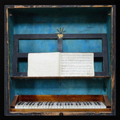 A vintage keyboard instrument is displayed inside a rustic wooden box with a distressed blue interior. An open book of sheet music rests on a black stand above the white and black keys. The entire piece is set within a simple, light-colored wood frame. Decor