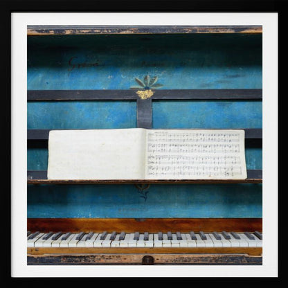 A vintage keyboard instrument is displayed inside a rustic wooden box with a distressed blue interior. An open book of sheet music rests on a black stand above the white and black keys. The entire piece is set within a simple, light-colored wood frame. Decor