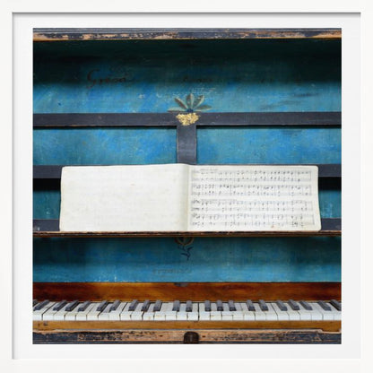 A vintage keyboard instrument is displayed inside a rustic wooden box with a distressed blue interior. An open book of sheet music rests on a black stand above the white and black keys. The entire piece is set within a simple, light-colored wood frame. Decor