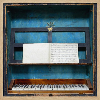A vintage keyboard instrument is displayed inside a rustic wooden box with a distressed blue interior. An open book of sheet music rests on a black stand above the white and black keys. The entire piece is set within a simple, light-colored wood frame. Decor