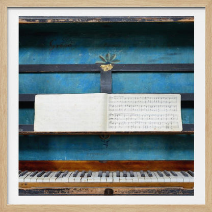 A vintage keyboard instrument is displayed inside a rustic wooden box with a distressed blue interior. An open book of sheet music rests on a black stand above the white and black keys. The entire piece is set within a simple, light-colored wood frame. Decor