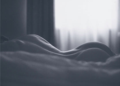 A close-up black and white artistic photograph capturing the soft curves of a person's back and hips as they lie down. The background is softly lit by a window, creating a play of light and shadow that emphasizes the human form's abstract, sculptural qualities. The image is displayed in a silver frame. Print