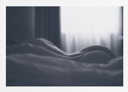 A close-up black and white artistic photograph capturing the soft curves of a person's back and hips as they lie down. The background is softly lit by a window, creating a play of light and shadow that emphasizes the human form's abstract, sculptural qualities. The image is displayed in a silver frame. Print