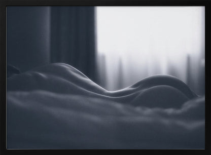 A close-up black and white artistic photograph capturing the soft curves of a person's back and hips as they lie down. The background is softly lit by a window, creating a play of light and shadow that emphasizes the human form's abstract, sculptural qualities. The image is displayed in a silver frame. Print