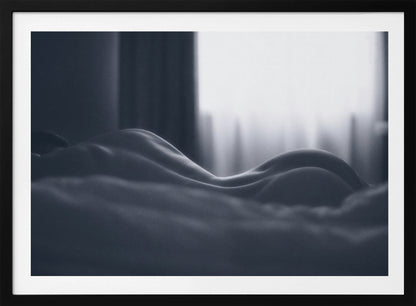 A close-up black and white artistic photograph capturing the soft curves of a person's back and hips as they lie down. The background is softly lit by a window, creating a play of light and shadow that emphasizes the human form's abstract, sculptural qualities. The image is displayed in a silver frame. Print