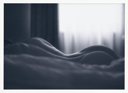 A close-up black and white artistic photograph capturing the soft curves of a person's back and hips as they lie down. The background is softly lit by a window, creating a play of light and shadow that emphasizes the human form's abstract, sculptural qualities. The image is displayed in a silver frame. Print