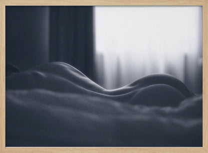 A close-up black and white artistic photograph capturing the soft curves of a person's back and hips as they lie down. The background is softly lit by a window, creating a play of light and shadow that emphasizes the human form's abstract, sculptural qualities. The image is displayed in a silver frame. Print