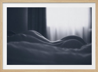 A close-up black and white artistic photograph capturing the soft curves of a person's back and hips as they lie down. The background is softly lit by a window, creating a play of light and shadow that emphasizes the human form's abstract, sculptural qualities. The image is displayed in a silver frame. Print