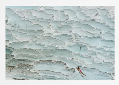 An aerial view of a woman in a pink bikini swimming in the stunning, terraced, milky-blue thermal pools of Pamukkale, Turkey. The white travertine formations create a unique, layered landscape, and the entire image is enclosed in a silver frame. Artwork