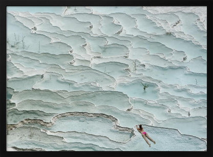 An aerial view of a woman in a pink bikini swimming in the stunning, terraced, milky-blue thermal pools of Pamukkale, Turkey. The white travertine formations create a unique, layered landscape, and the entire image is enclosed in a silver frame. Artwork