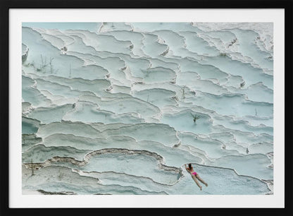 An aerial view of a woman in a pink bikini swimming in the stunning, terraced, milky-blue thermal pools of Pamukkale, Turkey. The white travertine formations create a unique, layered landscape, and the entire image is enclosed in a silver frame. Artwork