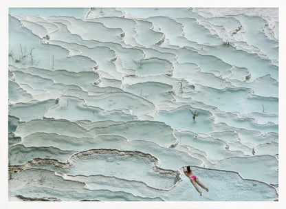 An aerial view of a woman in a pink bikini swimming in the stunning, terraced, milky-blue thermal pools of Pamukkale, Turkey. The white travertine formations create a unique, layered landscape, and the entire image is enclosed in a silver frame. Artwork
