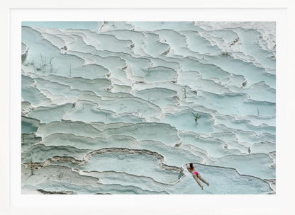 An aerial view of a woman in a pink bikini swimming in the stunning, terraced, milky-blue thermal pools of Pamukkale, Turkey. The white travertine formations create a unique, layered landscape, and the entire image is enclosed in a silver frame. Artwork