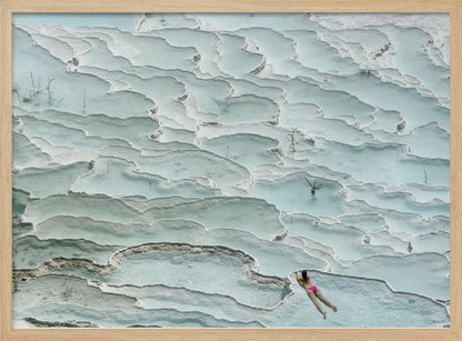 An aerial view of a woman in a pink bikini swimming in the stunning, terraced, milky-blue thermal pools of Pamukkale, Turkey. The white travertine formations create a unique, layered landscape, and the entire image is enclosed in a silver frame. Artwork