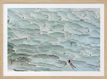 An aerial view of a woman in a pink bikini swimming in the stunning, terraced, milky-blue thermal pools of Pamukkale, Turkey. The white travertine formations create a unique, layered landscape, and the entire image is enclosed in a silver frame. Artwork
