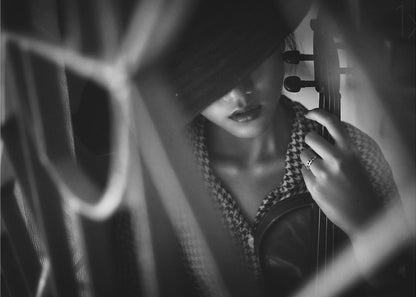 A moody, artistic black and white photograph of a woman holding a violin. Her face is partially obscured by a dark hat, creating a mysterious atmosphere. The image is shot through a blurred foreground, adding depth and intimacy to the portrait. Poster