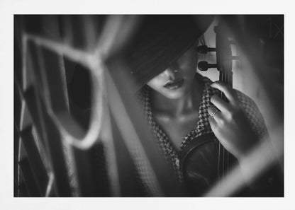 A moody, artistic black and white photograph of a woman holding a violin. Her face is partially obscured by a dark hat, creating a mysterious atmosphere. The image is shot through a blurred foreground, adding depth and intimacy to the portrait. Poster