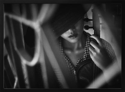 A moody, artistic black and white photograph of a woman holding a violin. Her face is partially obscured by a dark hat, creating a mysterious atmosphere. The image is shot through a blurred foreground, adding depth and intimacy to the portrait. Poster