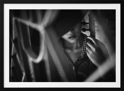 A moody, artistic black and white photograph of a woman holding a violin. Her face is partially obscured by a dark hat, creating a mysterious atmosphere. The image is shot through a blurred foreground, adding depth and intimacy to the portrait. Poster