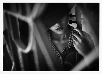 A moody, artistic black and white photograph of a woman holding a violin. Her face is partially obscured by a dark hat, creating a mysterious atmosphere. The image is shot through a blurred foreground, adding depth and intimacy to the portrait. Poster