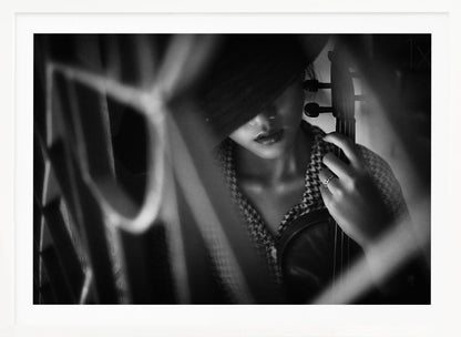 A moody, artistic black and white photograph of a woman holding a violin. Her face is partially obscured by a dark hat, creating a mysterious atmosphere. The image is shot through a blurred foreground, adding depth and intimacy to the portrait. Poster