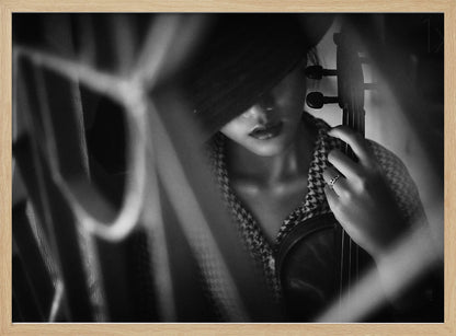 A moody, artistic black and white photograph of a woman holding a violin. Her face is partially obscured by a dark hat, creating a mysterious atmosphere. The image is shot through a blurred foreground, adding depth and intimacy to the portrait. Poster