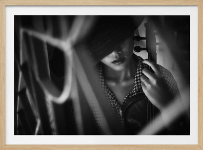 A moody, artistic black and white photograph of a woman holding a violin. Her face is partially obscured by a dark hat, creating a mysterious atmosphere. The image is shot through a blurred foreground, adding depth and intimacy to the portrait. Poster