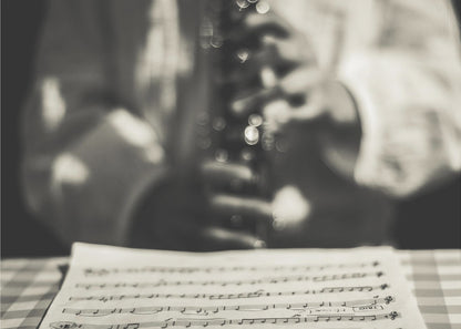 A shallow depth of field, black and white photograph focusing on sheet music in the foreground. The background is softly blurred, showing the hands of a musician holding a woodwind instrument. Wall Art