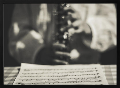 A shallow depth of field, black and white photograph focusing on sheet music in the foreground. The background is softly blurred, showing the hands of a musician holding a woodwind instrument. Wall Art