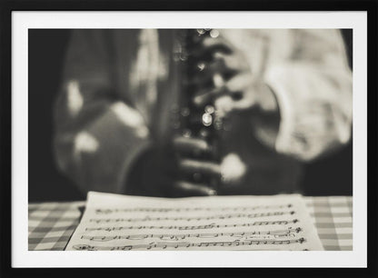 A shallow depth of field, black and white photograph focusing on sheet music in the foreground. The background is softly blurred, showing the hands of a musician holding a woodwind instrument. Wall Art