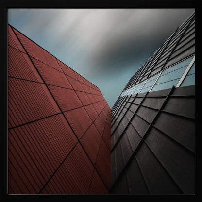 A dramatic low-angle photograph of two modern buildings with contrasting facades. On the left, a building is covered in deep red square panels. On the right, a darker building features black angular tiles and glass windows. The sky above is a moody, blurred grey. Print