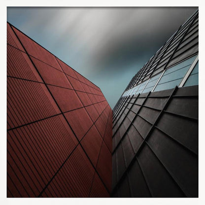 A dramatic low-angle photograph of two modern buildings with contrasting facades. On the left, a building is covered in deep red square panels. On the right, a darker building features black angular tiles and glass windows. The sky above is a moody, blurred grey. Print